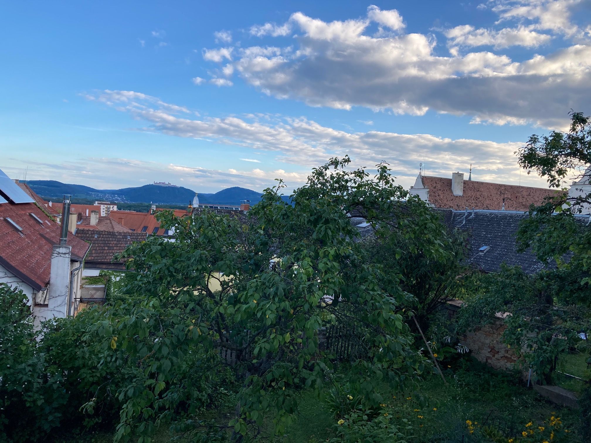 View over roofs and trees to hills and blue sky with clouds.
