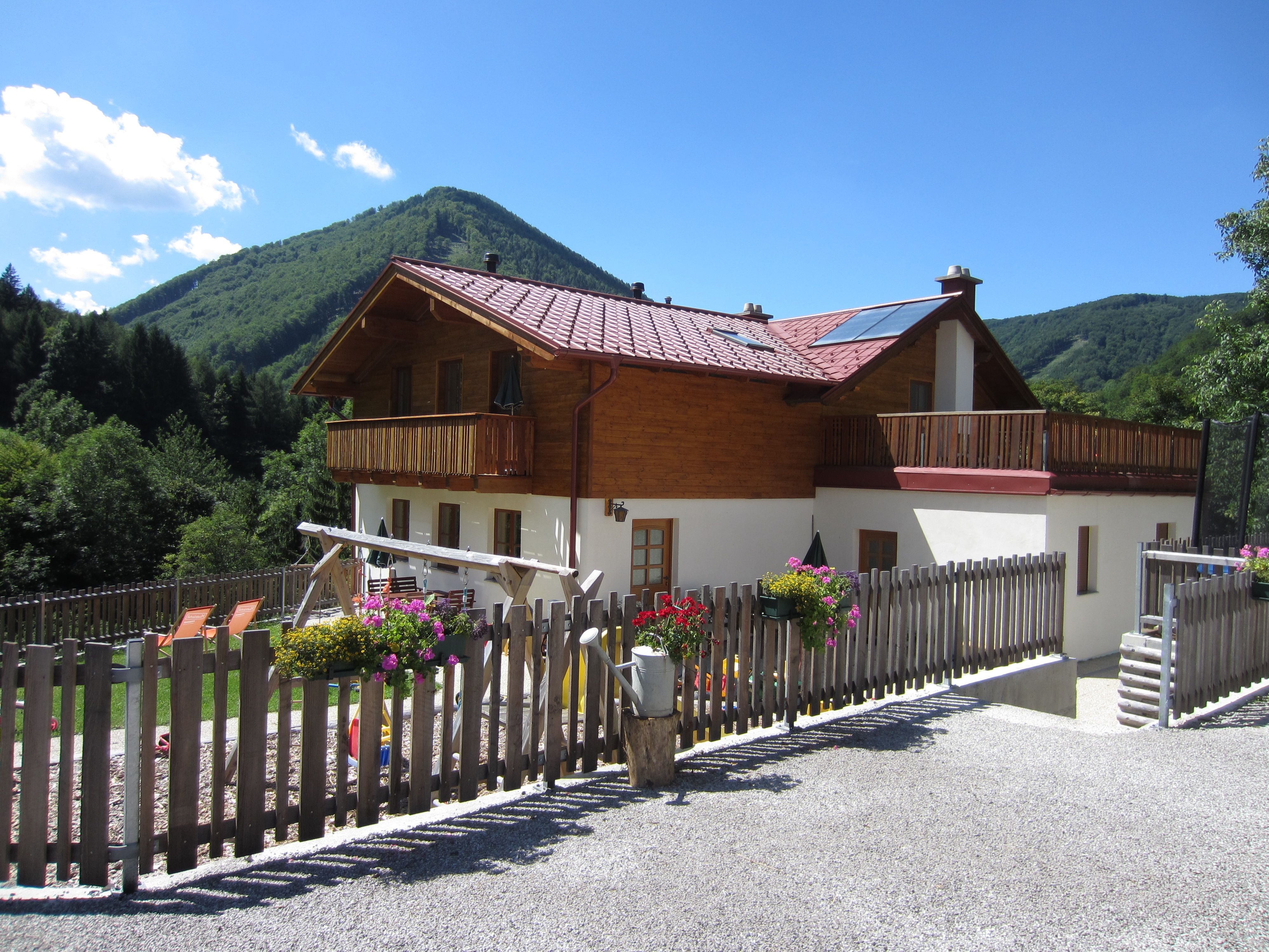 A two-storey house with a wooden balcony and garden, surrounded by mountains and trees.