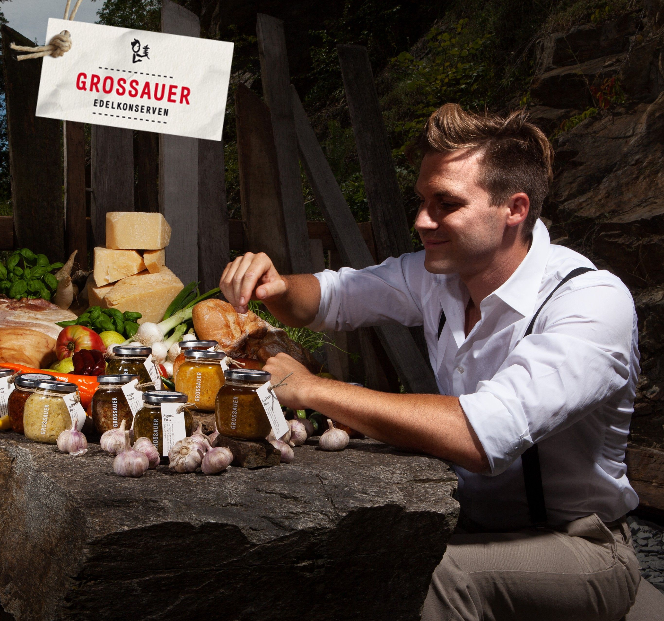 Man arranges jars of food on an outdoor table, surrounded by cheese, vegetables and bread.