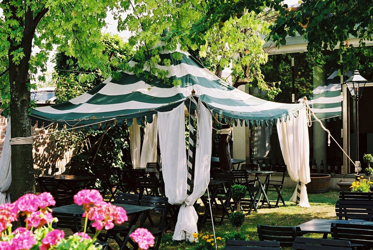 A striped tent with white curtains stands in a garden surrounded by trees and flowers.