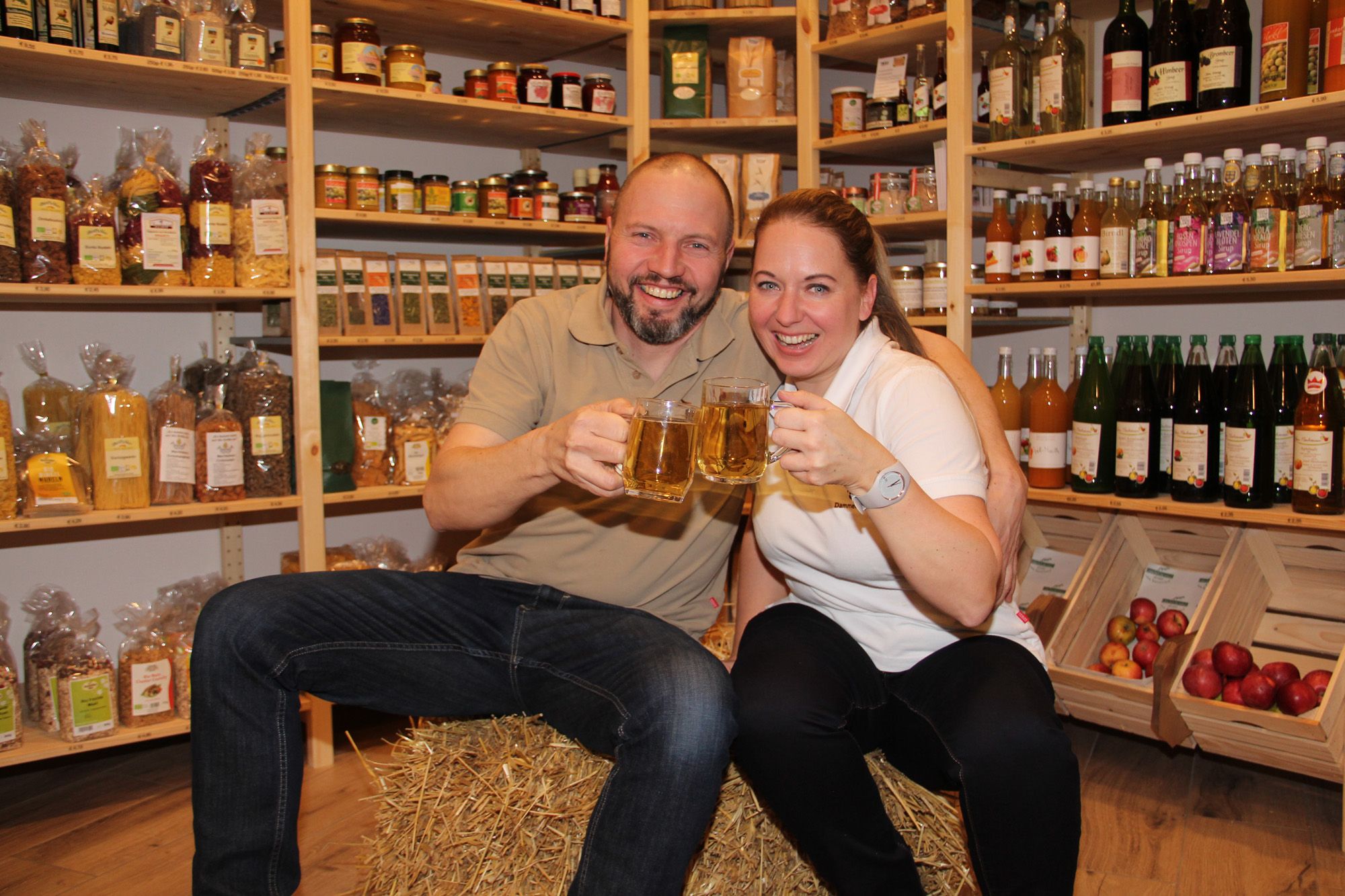 Two people sit on a hay bale in the farm store and toast with drinks.