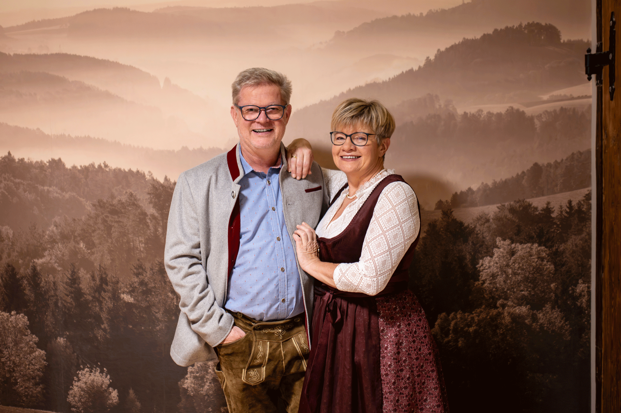 A couple in traditional Bavarian dress stands in front of a background with a forest landscape.