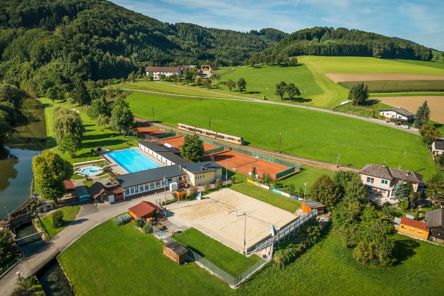 Aerial view of an outdoor swimming pool with pool, tennis courts and volleyball court, surrounded by green fields and hills.