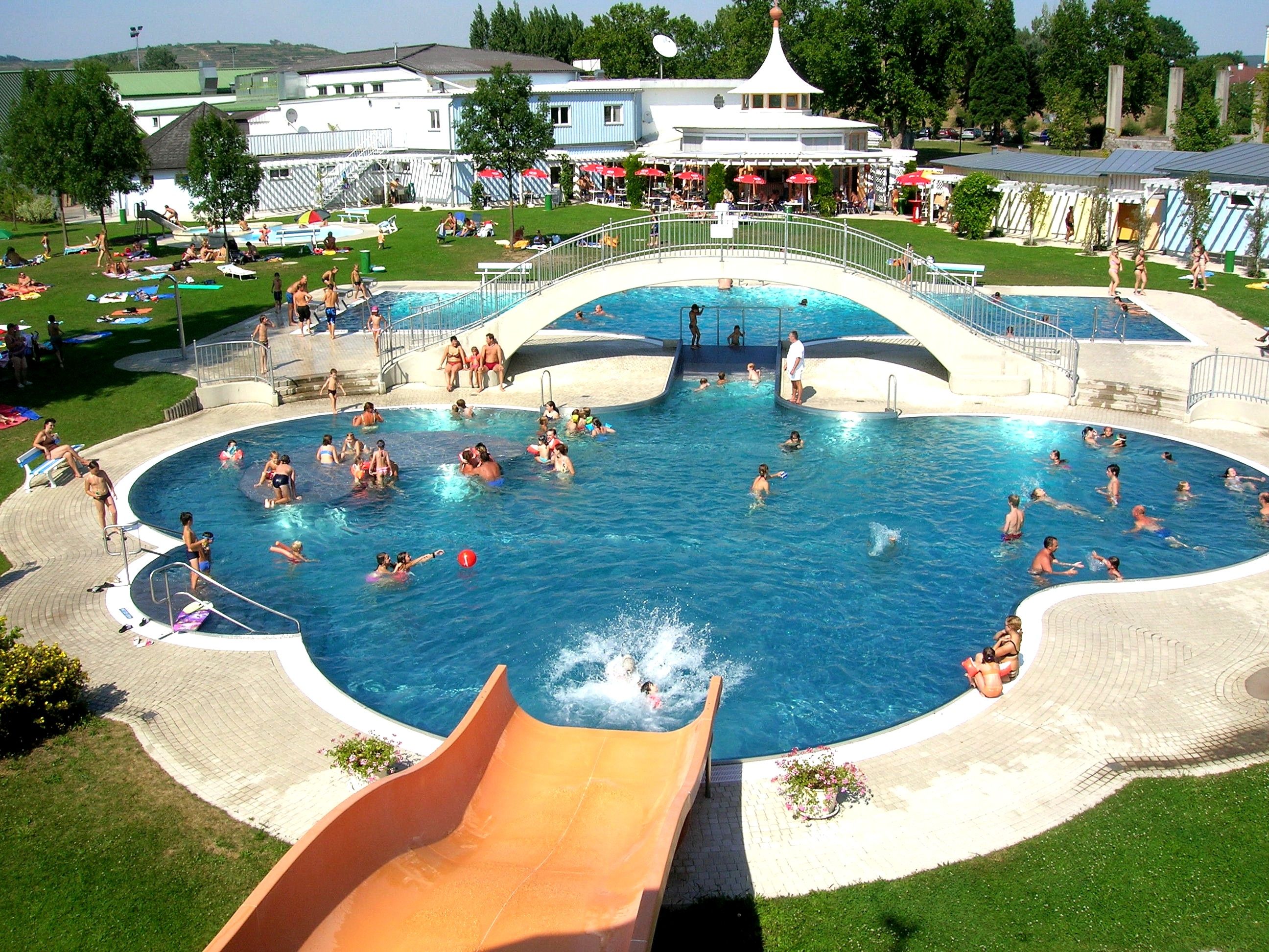 Outdoor swimming pool with bridge and slide, lots of people swimming and sunbathing.