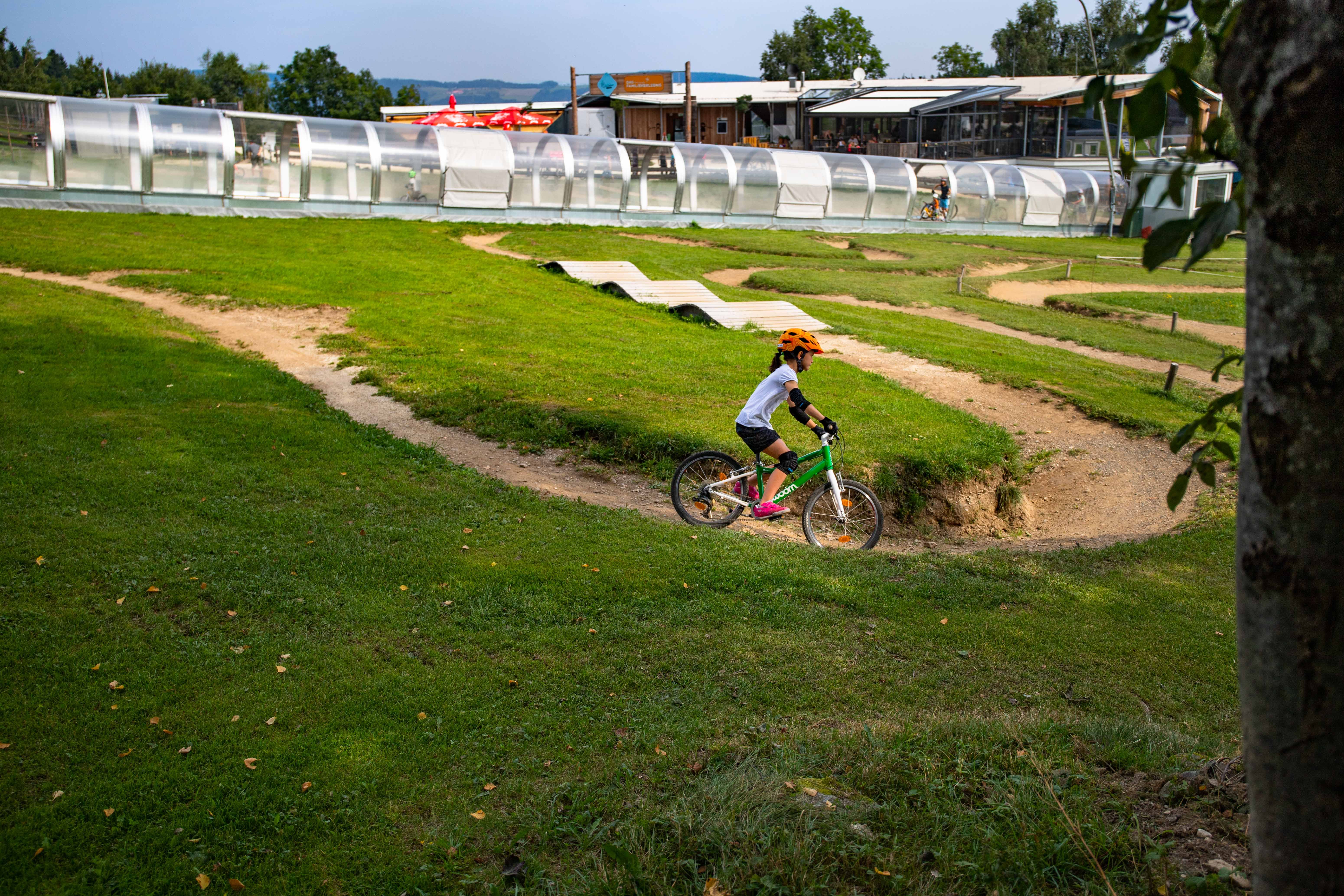 A child with an orange helmet rides a green bike through a mini bike park with hills and bends.