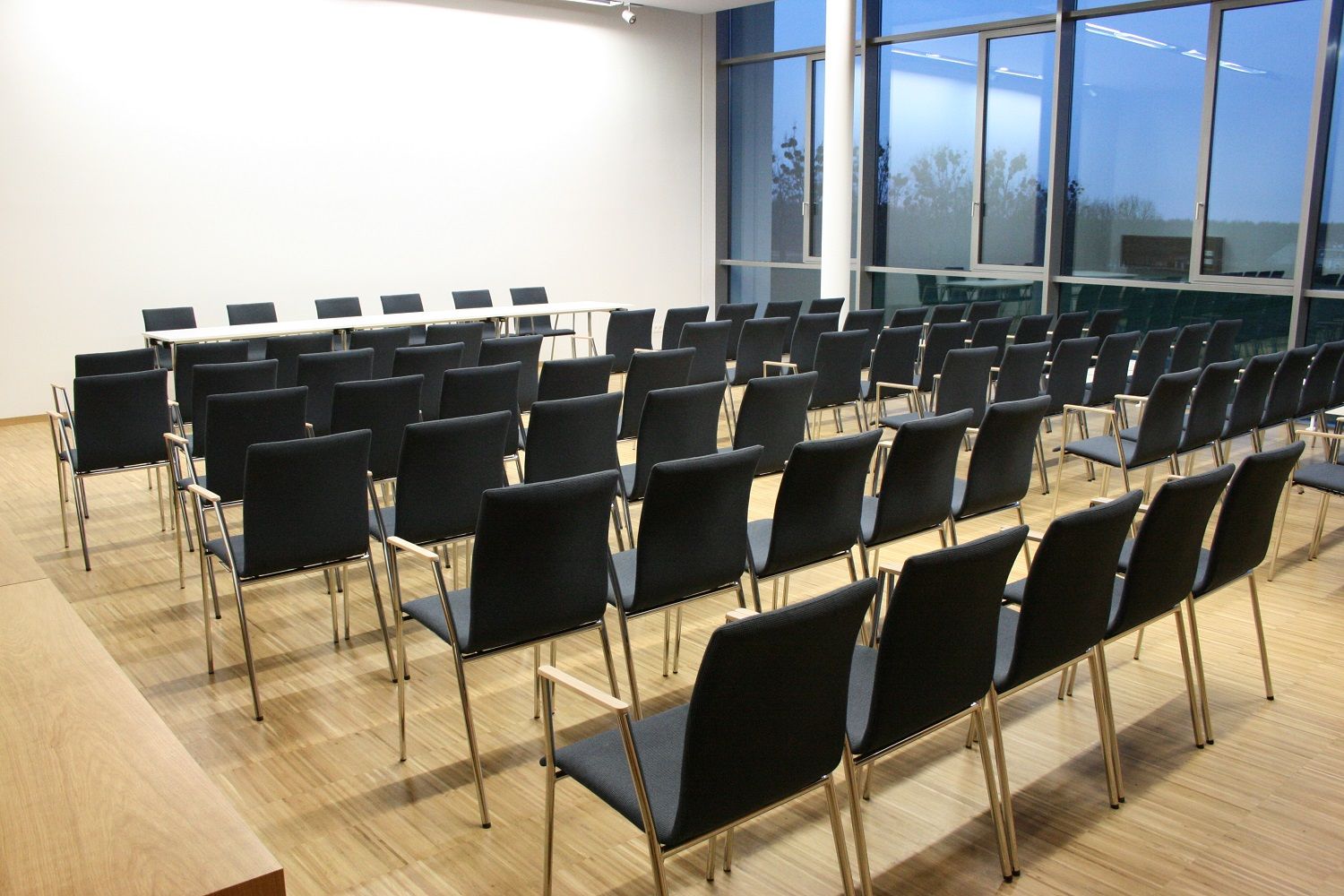 An empty seminar room with several rows of black chairs and a table at the front, large windows at the side.