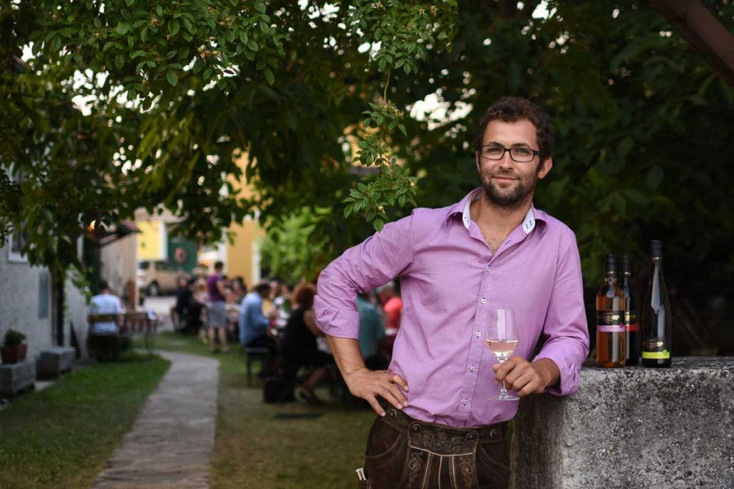 Man in purple shirt with wine glass, standing outside next to bottles.