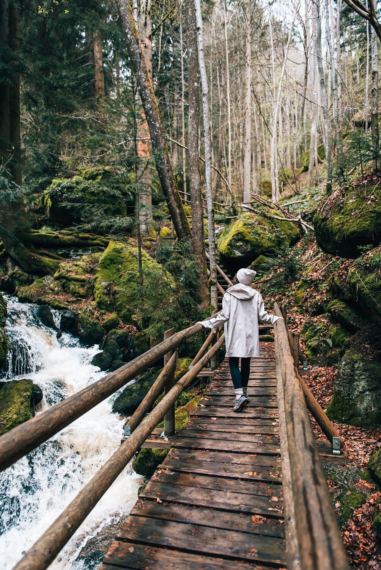 Ein sanfter Wasserfall plätschert über die moosbedeckten Steine, während Wanderer auf der Holzbrücke die Ruhe der Natur genießen. Umgeben von hohen Bäumen und frischem Grün, lädt dieser Ort dazu ein, die Seele baumeln zu lassen und die Schönheit der Umgebung zu erkunden.