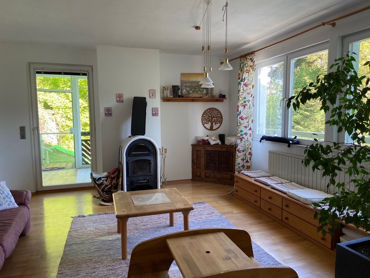 A cozy living room with wood-burning stove, wooden furniture and large windows with a view of the greenery.