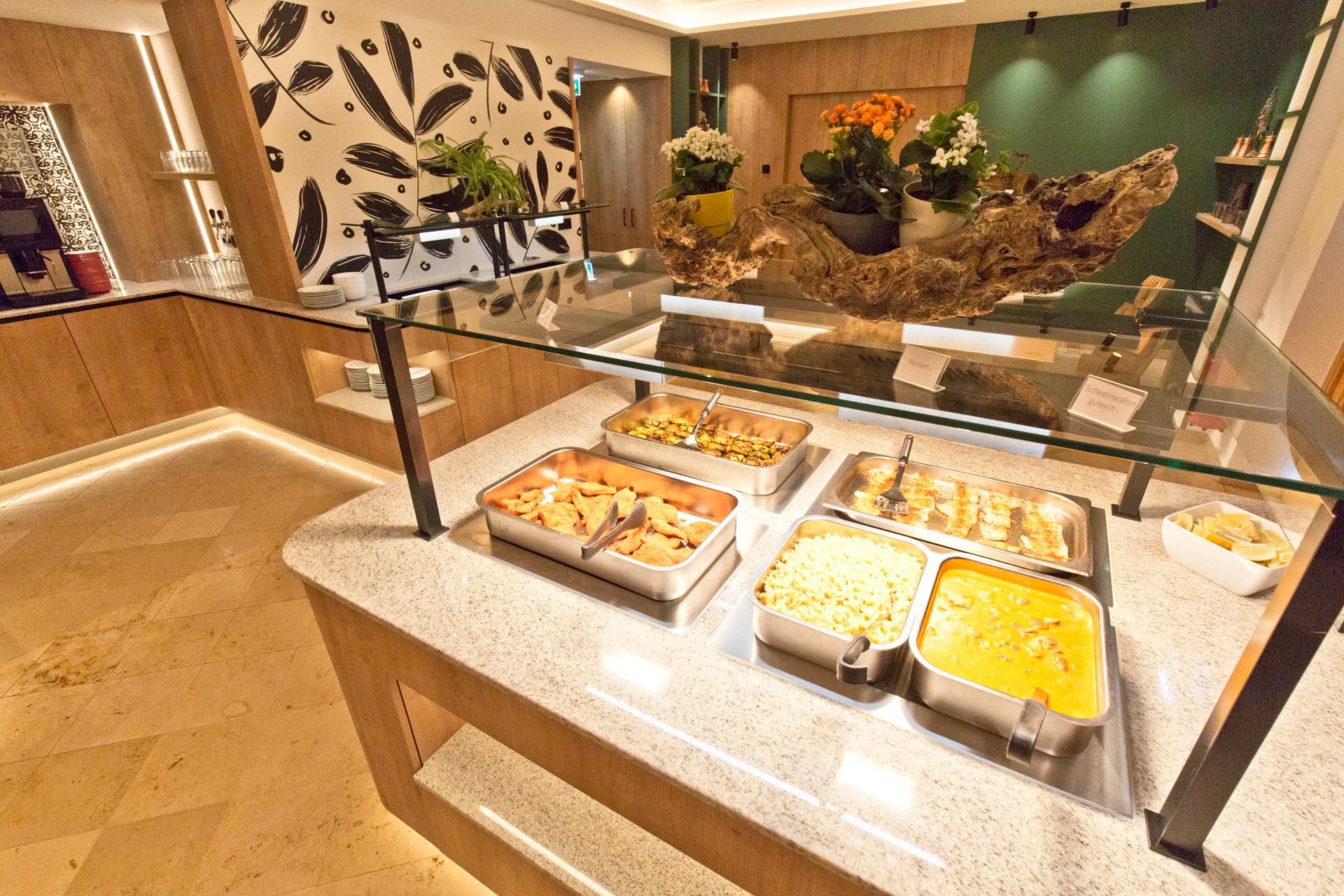 Buffet area with various dishes in stainless steel containers, decorated with plants and wooden elements.