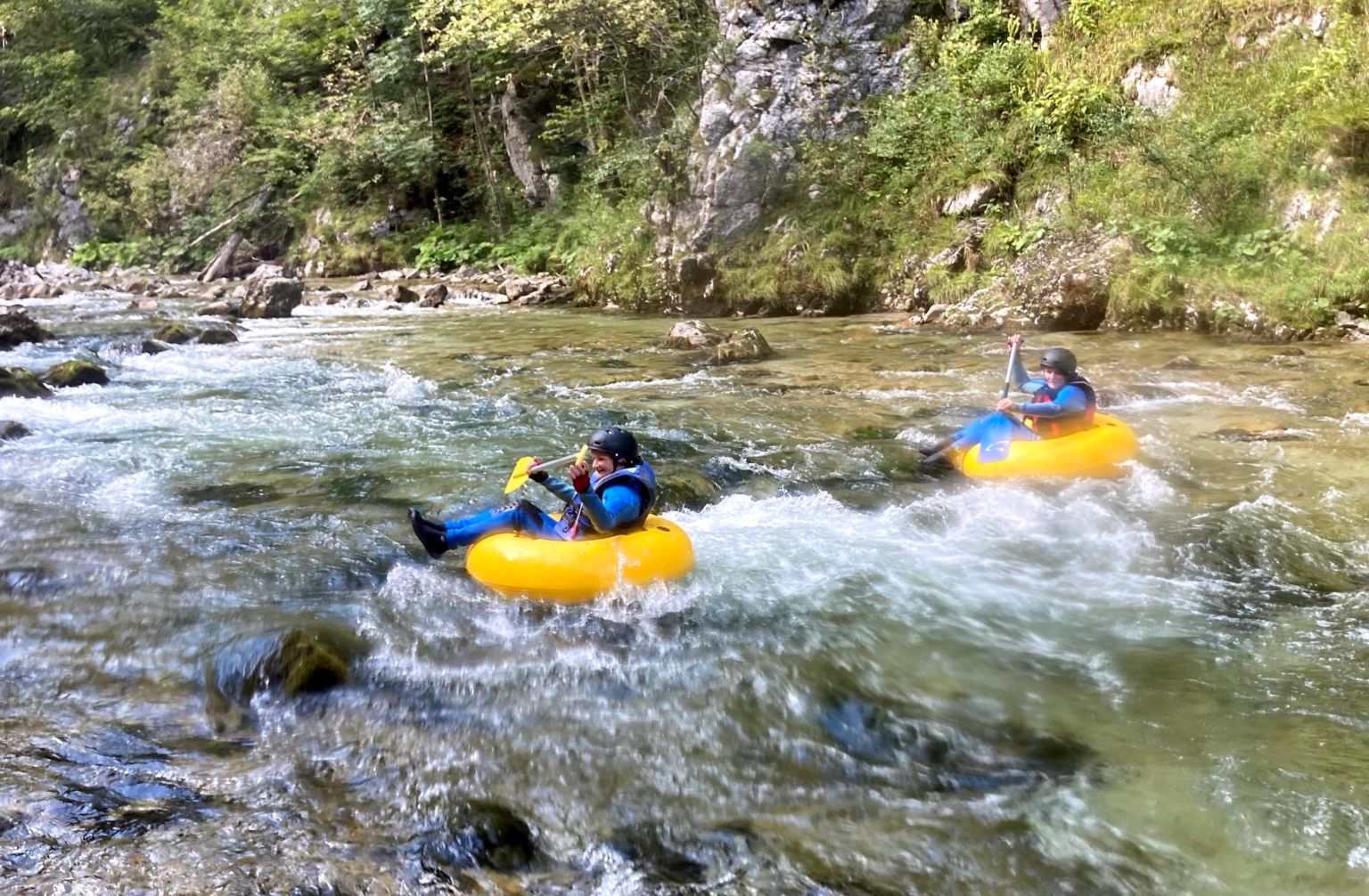 Two people tubing on a river in a wooded area.