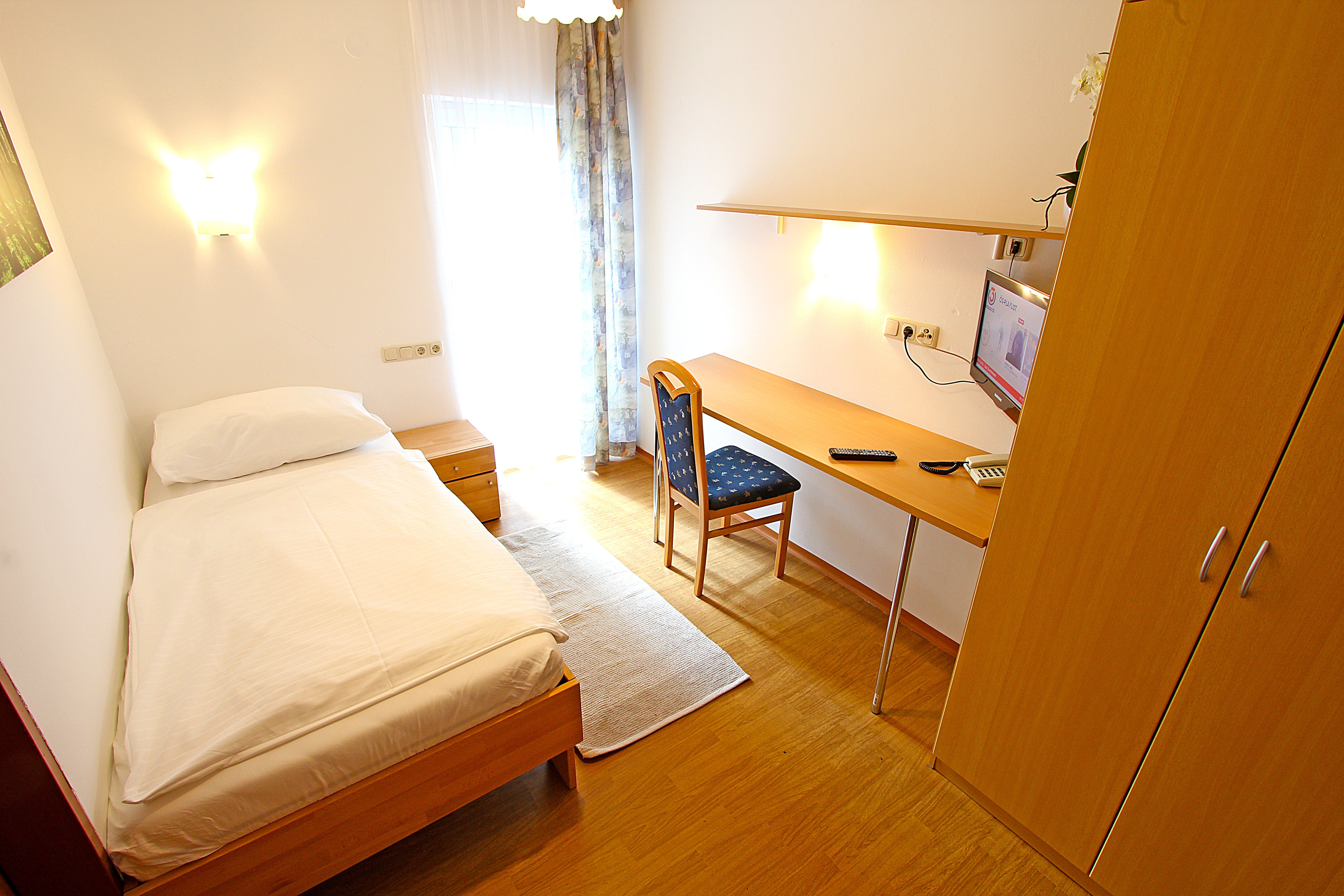 A simply furnished single room with bed, desk, chair and TV.