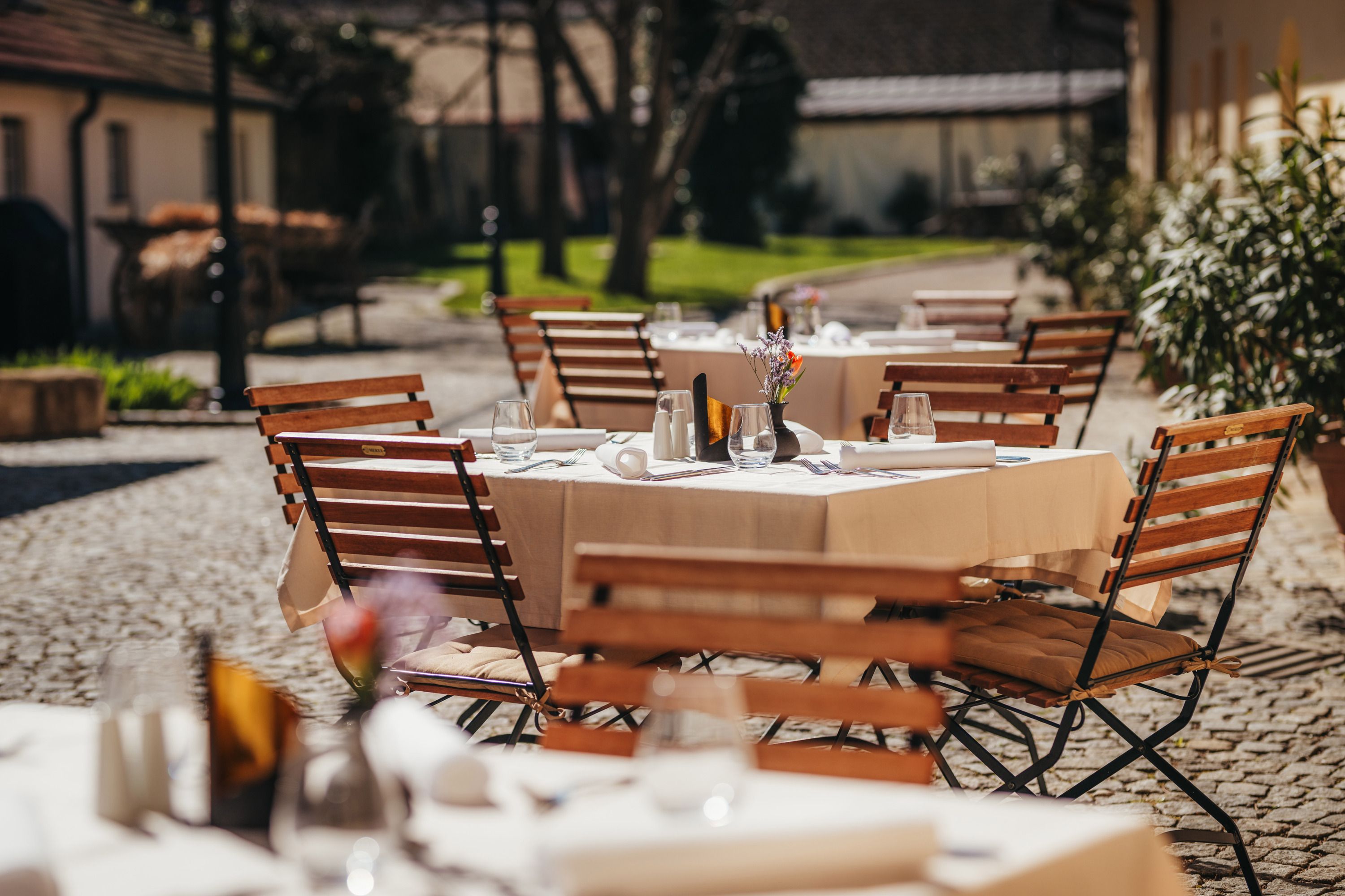 A sunny guest garden with wooden tables and chairs, set with white tablecloths and glasses, in a paved courtyard.