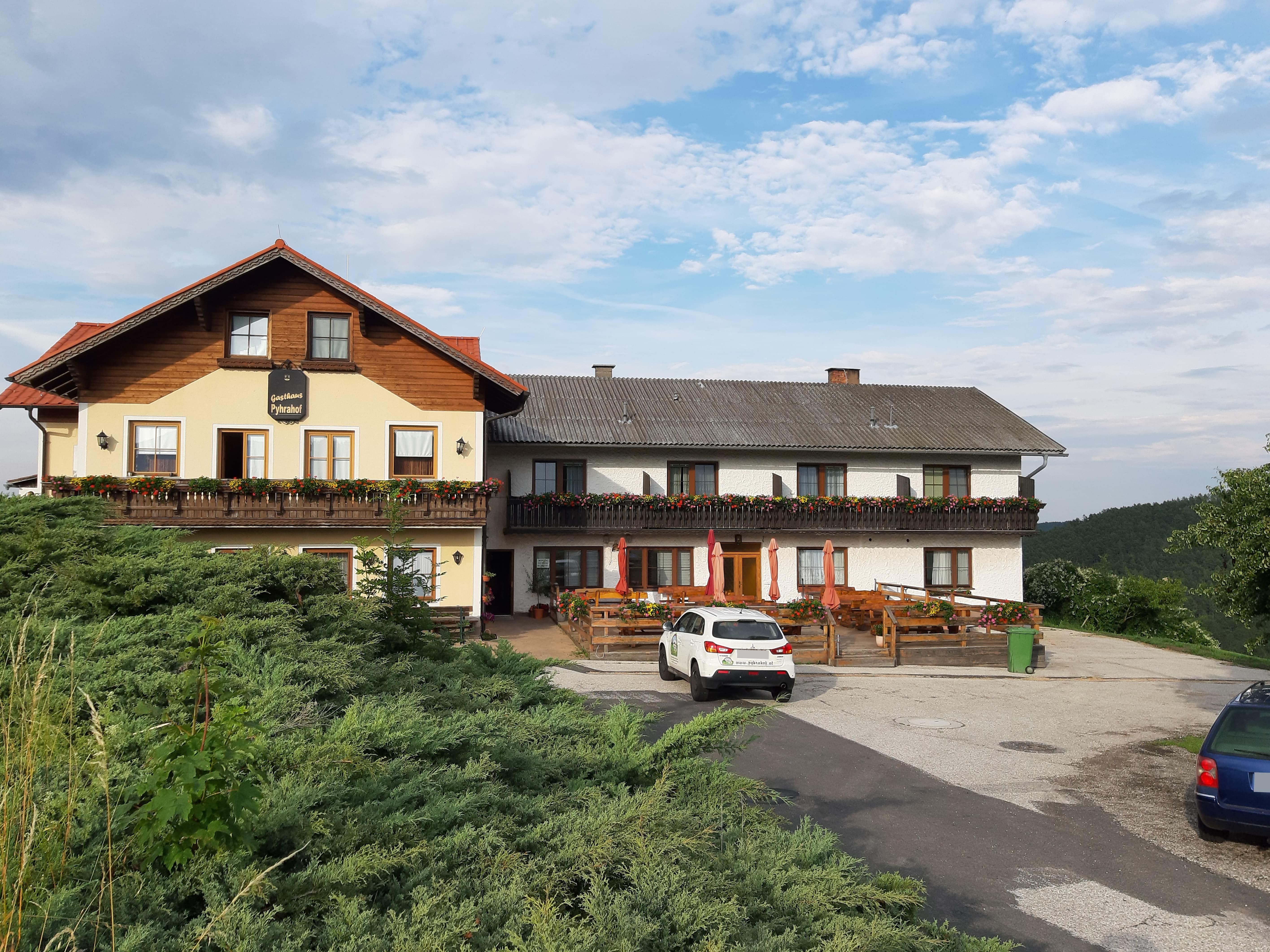 A traditional inn with a balcony and flower boxes, a parking lot with cars in front, surrounded by a green landscape and blue sky.