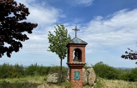 A brick wayside shrine with a cross on a hill, surrounded by trees and bushes, under a blue sky.