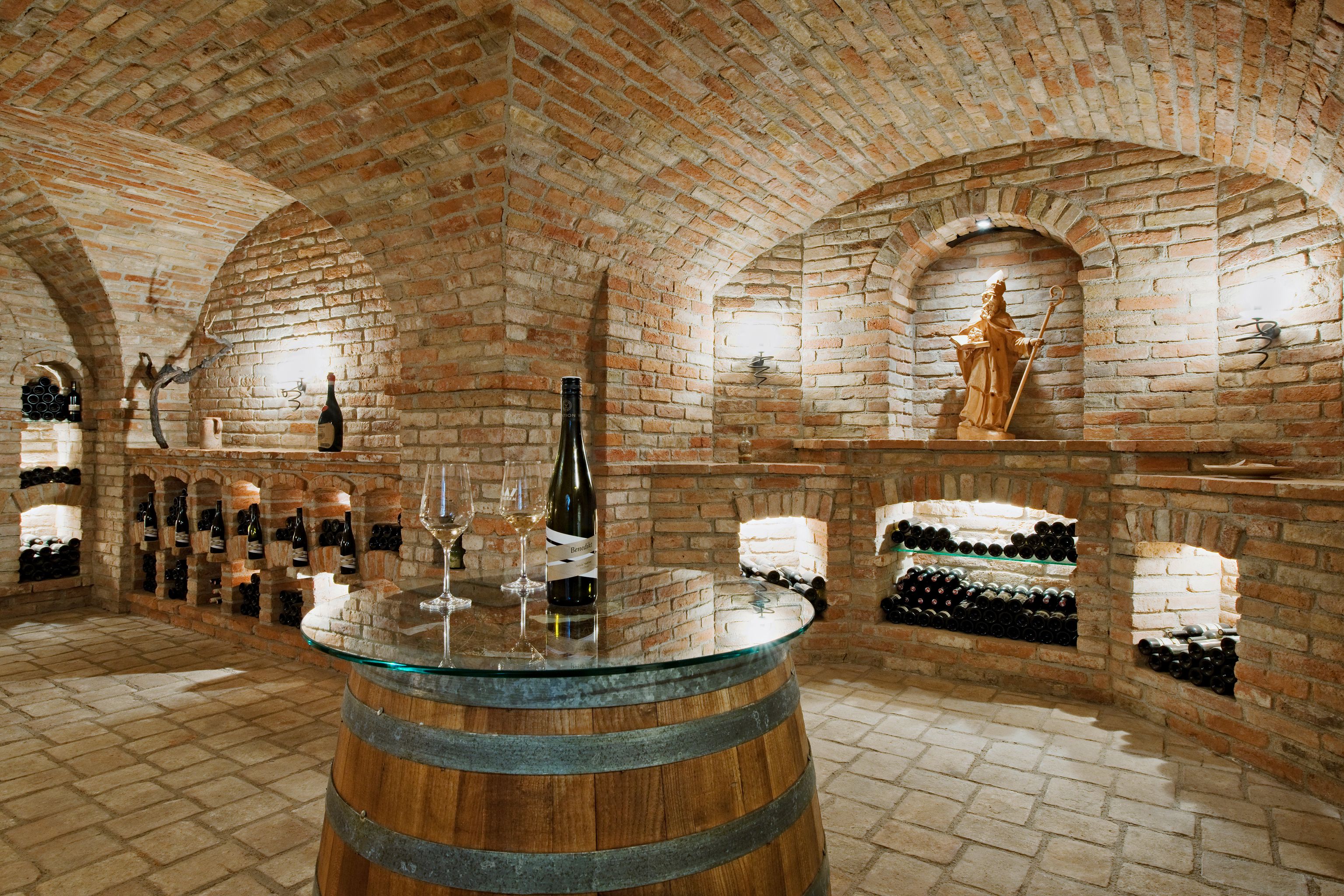 A vaulted cellar with brick walls, wine bottles on shelves and a barrel with a glass top as a table.