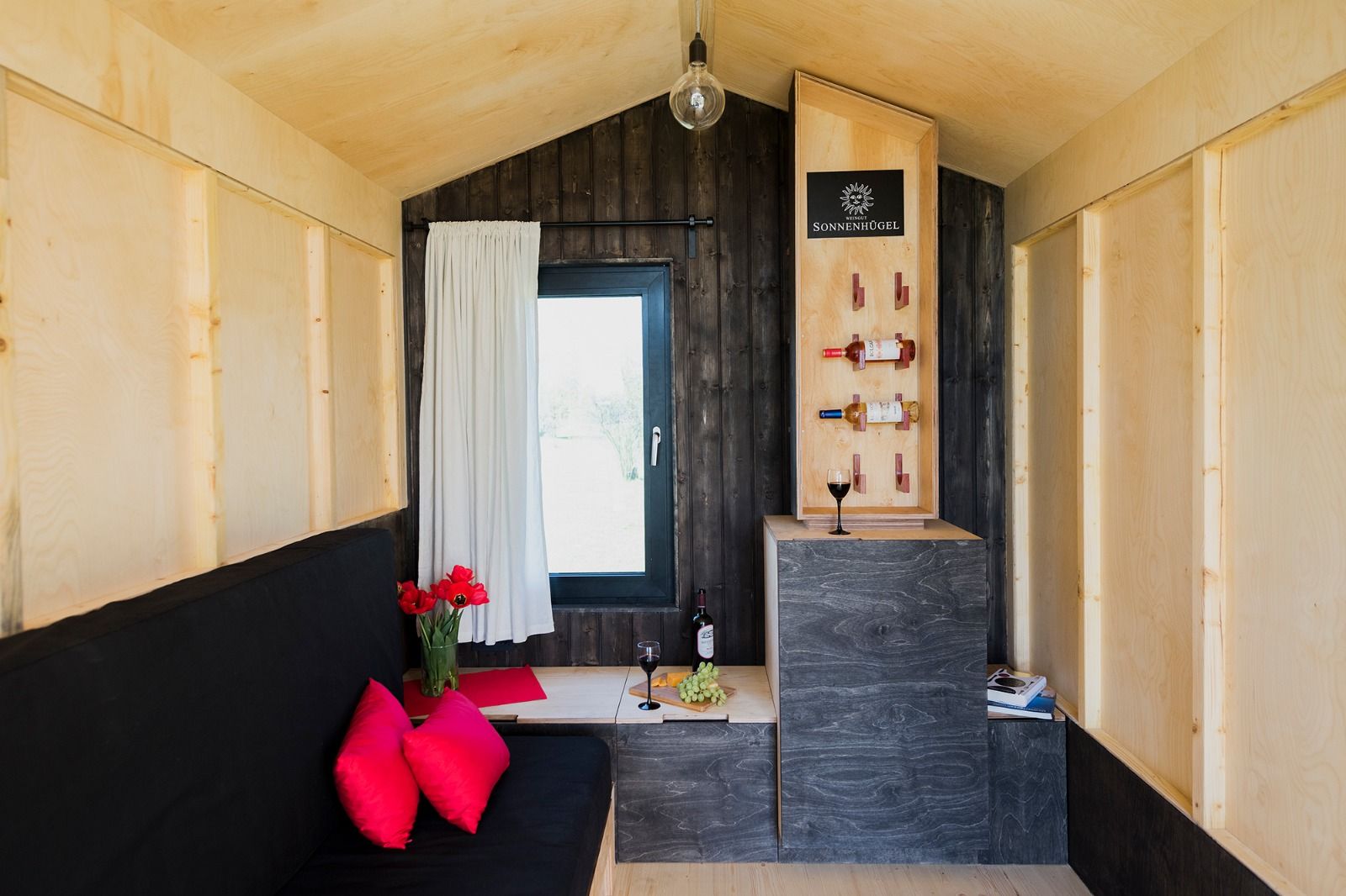 Interior view of a cozy vineyard hut with wooden walls, a sofa, red cushions, a wine glass and wine bottles.