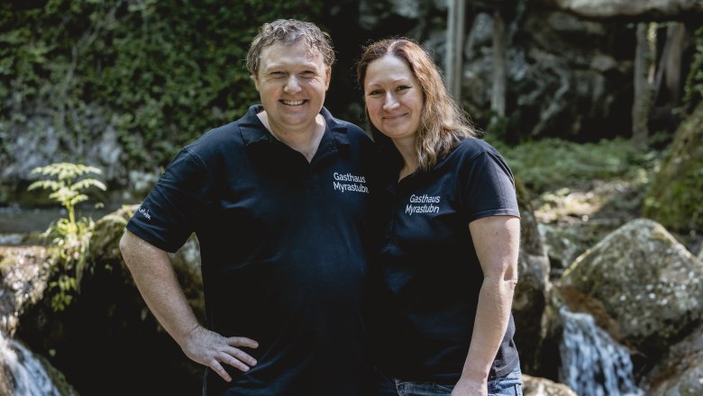 A man and a woman in black T-shirts with the inscription 'Gasthaus Myrastubn' stand smiling in front of a waterfall.