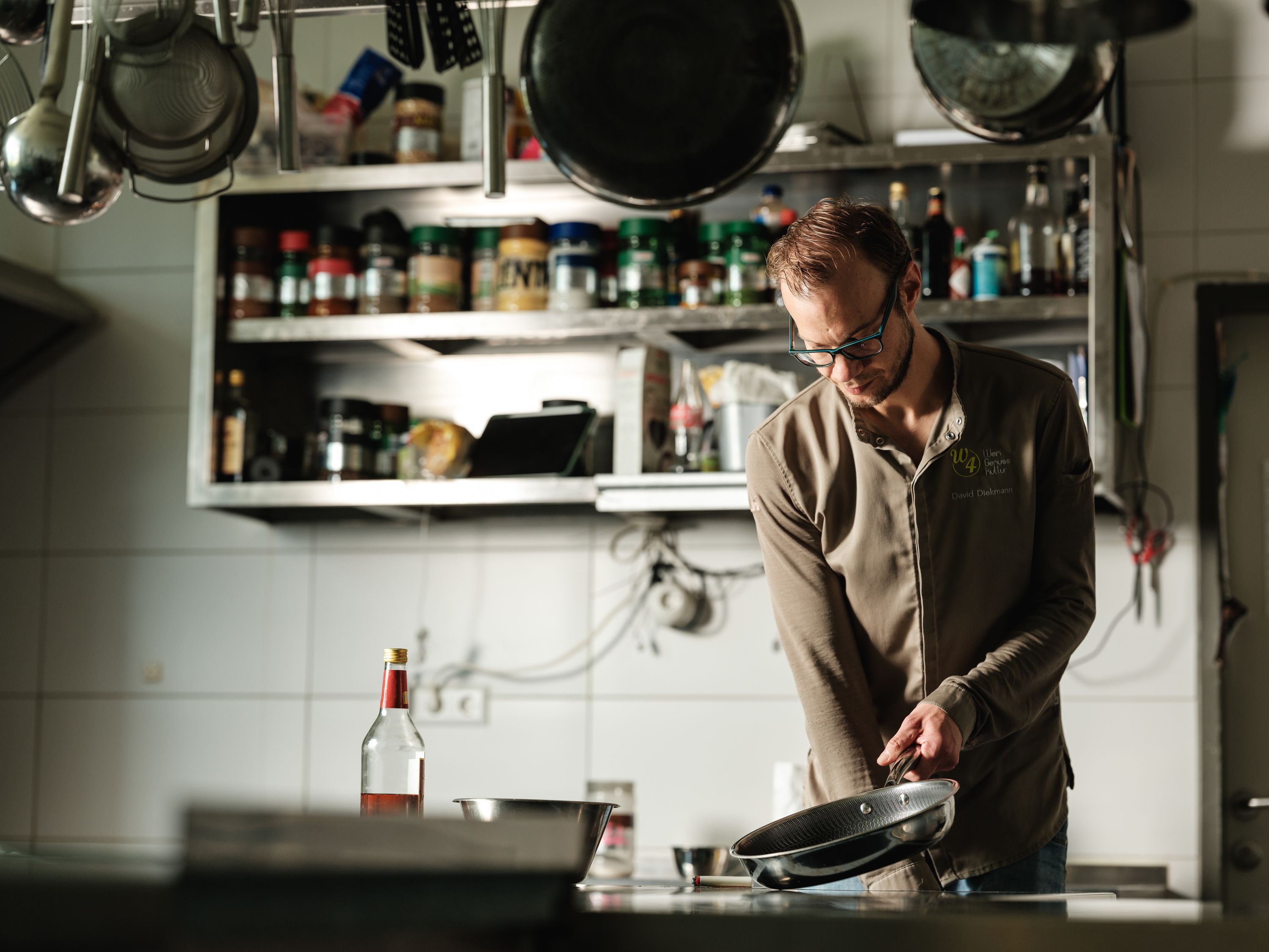 A chef in a professional kitchen holds a pan. Spice racks and kitchen utensils can be seen in the background.