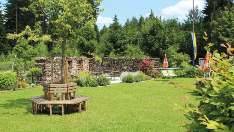 A green garden with wooden benches around a tree, surrounded by woods and flowers.