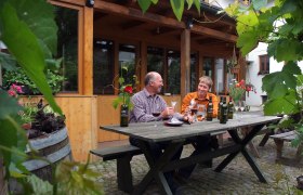 Two men sit at a wooden table outside and taste wine. Surrounded by plants and a wooden building in the background.