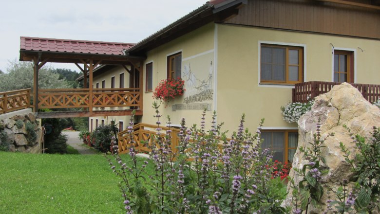 Stone builder, © Fam. König A yellow building with a wooden balcony and flowers, surrounded by green countryside.