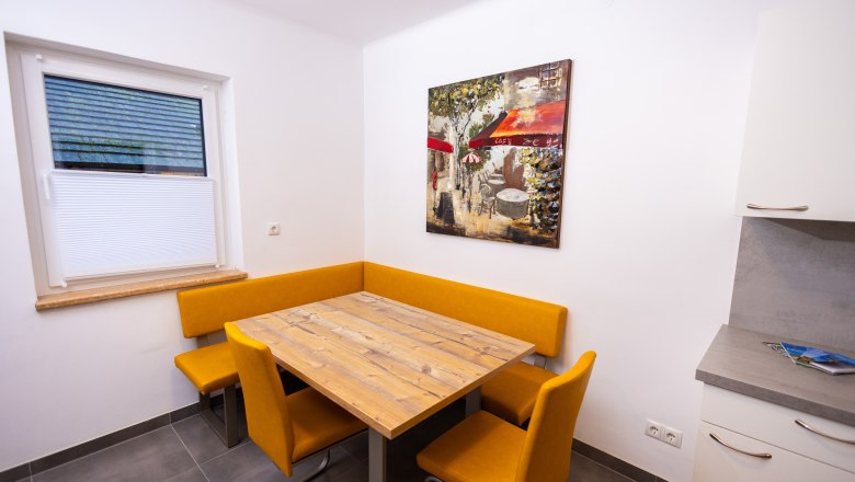 Dining area with wooden table, yellow corner bench and chairs, picture on the wall.