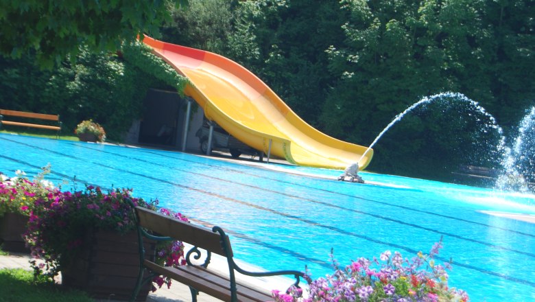 An outdoor pool with a yellow slide, water fountain and flowers in the foreground.