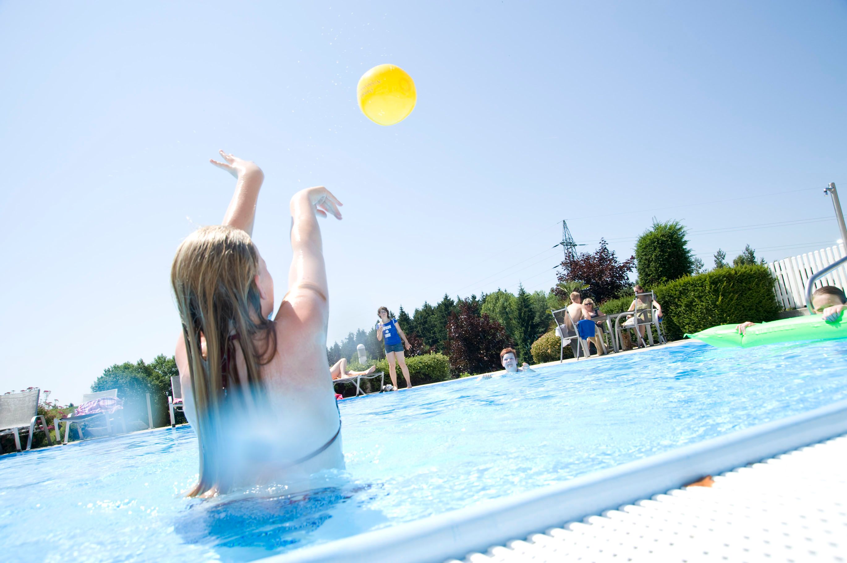 Person in pool throwing yellow ball, surrounded by other people and trees.