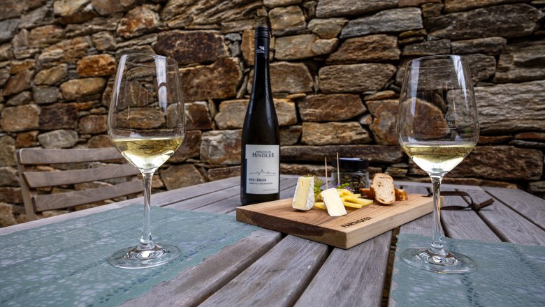 Two wine glasses and a bottle of wine on a wooden table with a cheese board, in front of a stone wall.