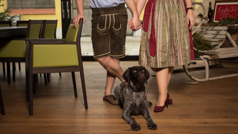 A couple in traditional dress hold hands, a dog sits at their feet.