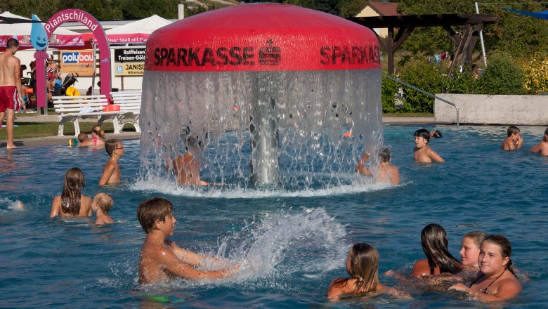 People swimming in an outdoor pool with a large water bubbler in the middle.
