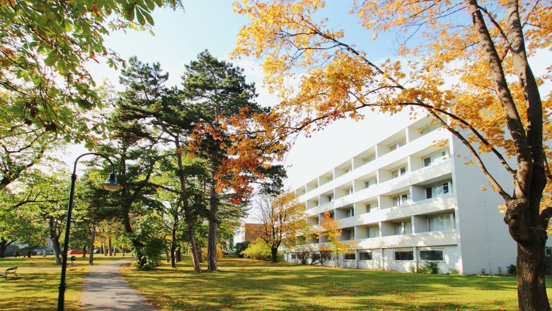 Surrounded by the black pine forest in Bad Vöslau, © College Garden Hotel A white building next to an autumnal park with trees and a sidewalk in Bad Vöslau.