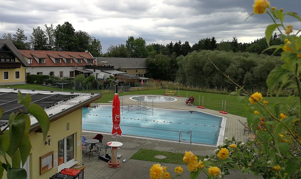 An outdoor swimming pool, surrounded by buildings and trees, under a cloudy sky.