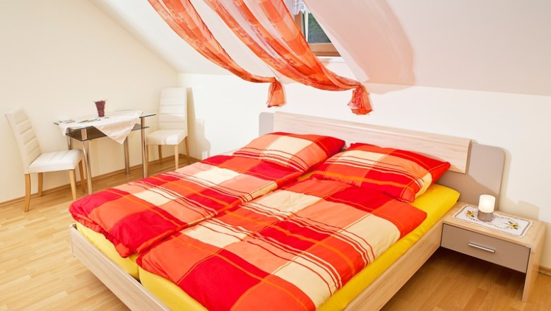 Room Pehn 2, © Gerhard Pehn Bright bedroom with red comforter cover and decorative curtains.