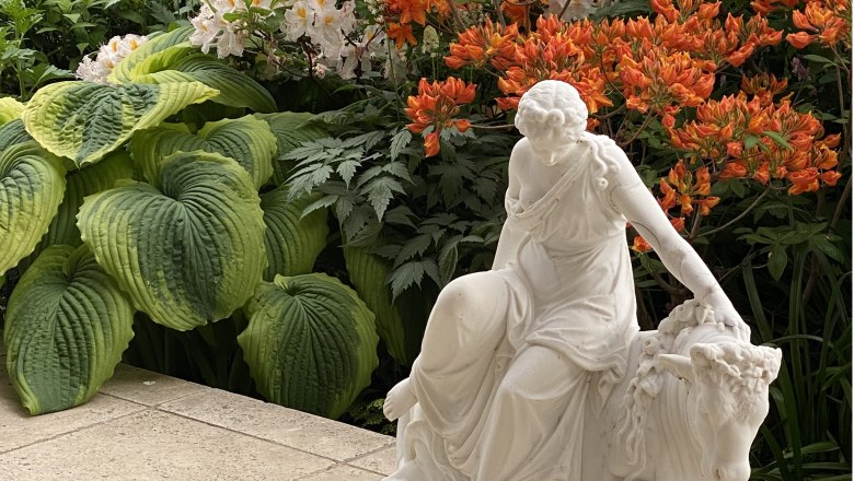 White statue of a woman next to large green leaves and orange flowers.