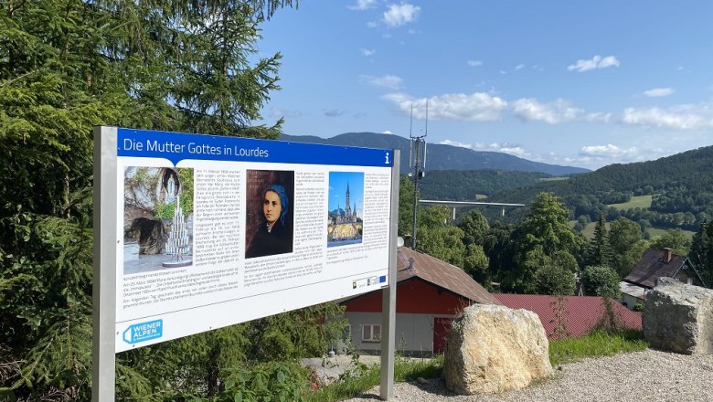 Information sign about the Mother of God in Lourdes in front of a landscape with hills and trees.