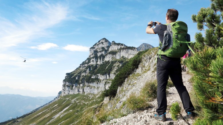 A hiker with a green rucksack photographs a mountain landscape with a helicopter in the sky.