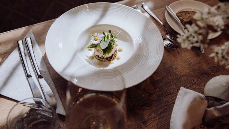 A plate of smoked trout tartare on a potato pancake, garnished with herbs and flowers, on a wooden table with cutlery and a glass of wine.
