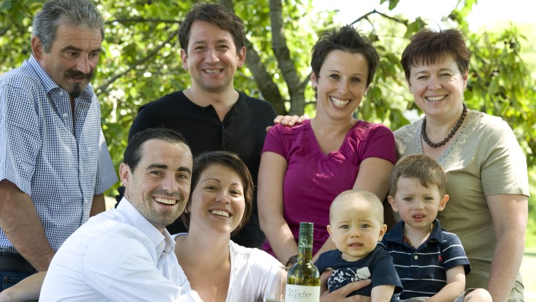 A smiling family poses outdoors with a bottle of wine on a table.