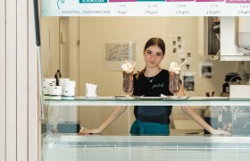 A woman stands behind an ice cream counter in an ice cream store, surrounded by different types of ice cream on a sign.