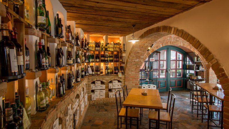 Wine cellar with shelves full of wine bottles, wooden tables and brick arches.