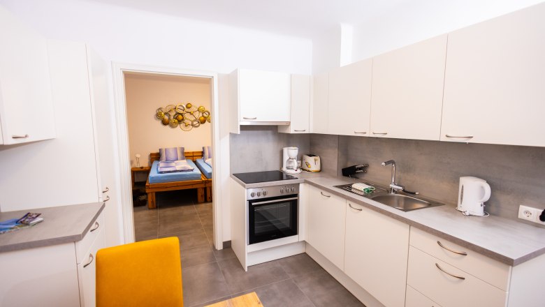 Modern kitchen with white cupboards, stove, sink and view into the bedroom.