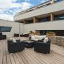 Modern terrace with wooden floor, black rattan furniture and stone wall, surrounded by a modern building.