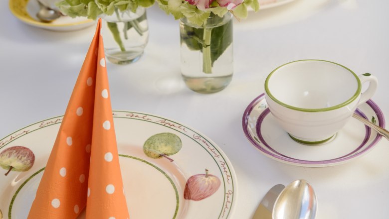 An elegantly laid table with flowers, orange dotted napkins and crockery with fruit motifs.