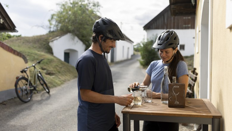 Two cyclists with helmets stand at a table in a wine cellar lane and pour themselves drinks.