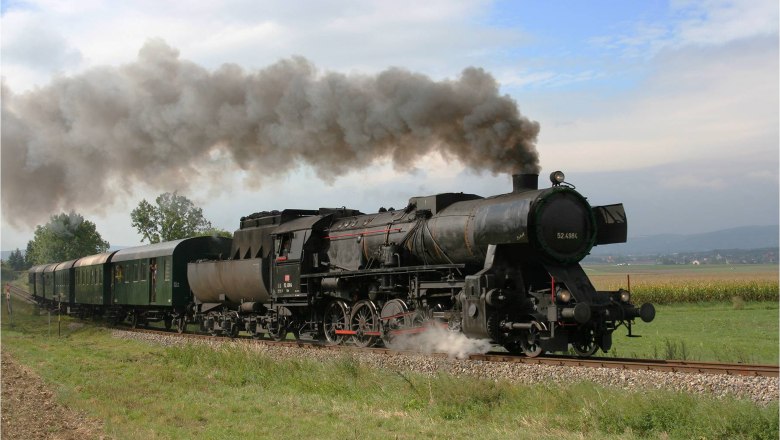 A steaming black steam locomotive pulls green wagons through a rural landscape.