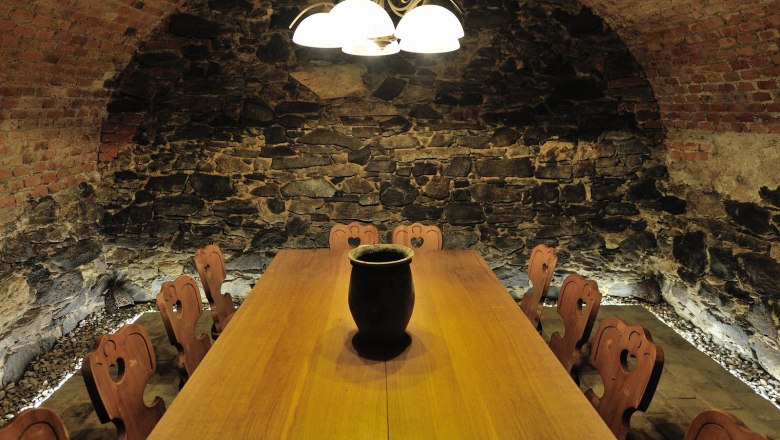 A rustic wine cellar with a long wooden table and chairs, surrounded by stone walls and illuminated by a ceiling lamp.