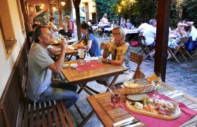 People sit at wooden tables in a cozy outdoor area and enjoy food and drinks.