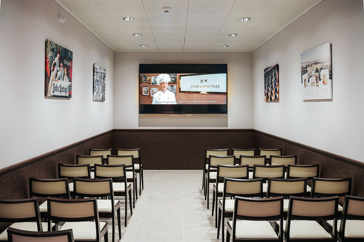 An empty lecture room with rows of chairs and a screen showing a presentation by Lindt & Sprüngli.